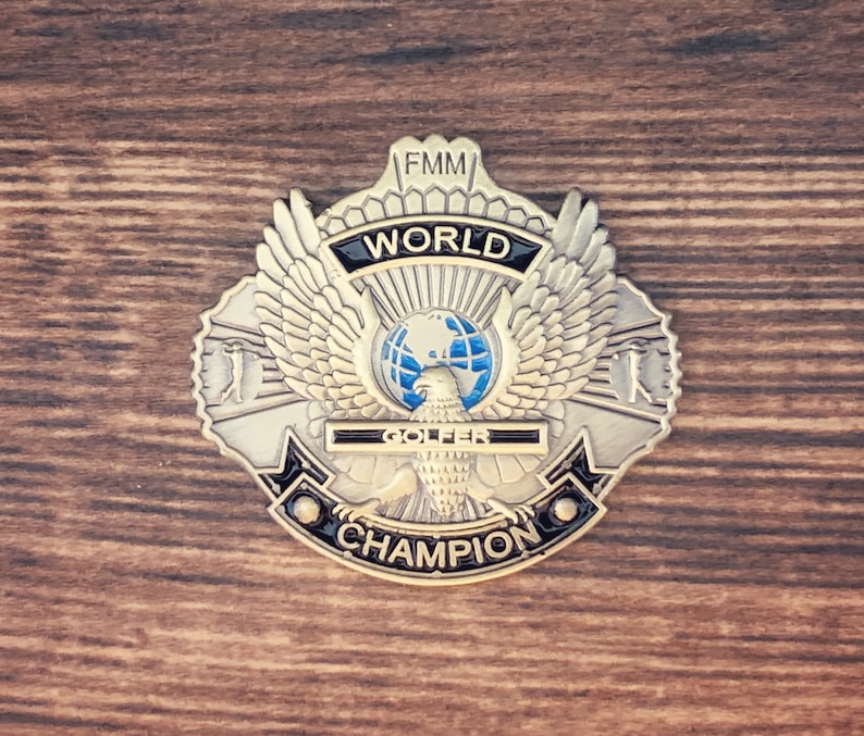 May include: A gold-colored metal badge with an eagle design and the text "FMM WORLD GOLFER CHAMPION".
