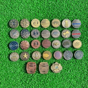 May include: A collection of 25 different golf ball markers. The markers are made of metal and feature a variety of designs, including flags, animals, and abstract patterns. The markers are arranged in a grid pattern on a green surface.