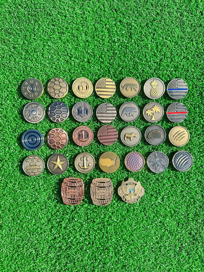 May include: A collection of 25 different golf ball markers. The markers are made of metal and feature a variety of designs, including flags, animals, and abstract patterns. The markers are arranged in a grid pattern on a green surface.