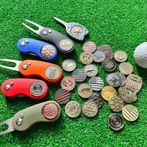 May include: A set of colorful golf ball markers with various designs, including a pineapple, a palm tree, and a star. The markers are arranged on a green surface with a golf ball and other golf accessories.