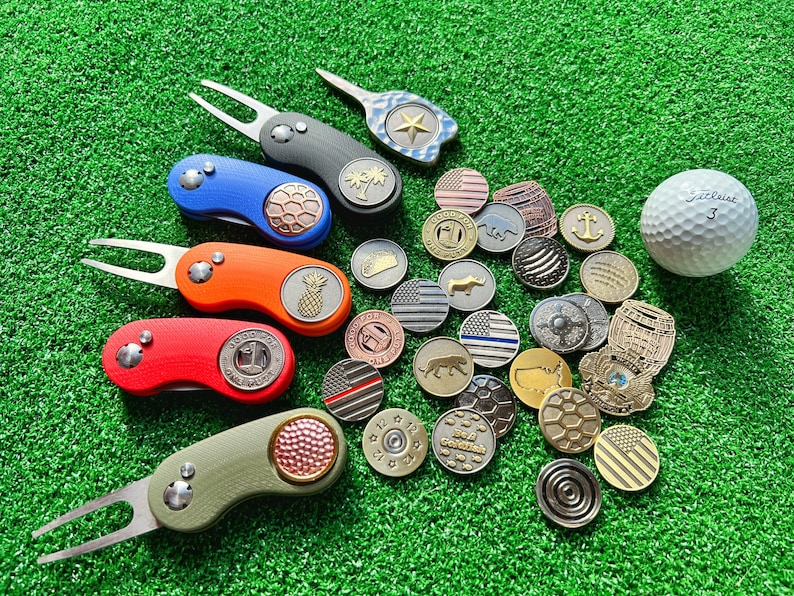May include: A set of colorful golf ball markers with various designs, including a pineapple, a palm tree, and a star. The markers are arranged on a green surface with a golf ball and other golf accessories.