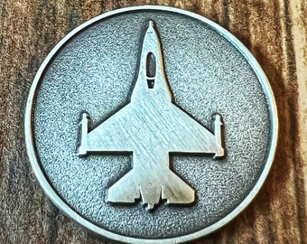 May include: A silver-colored metal coin with a detailed silhouette of a fighter jet in the center.