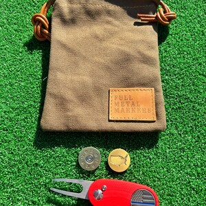 May include: A set of golf accessories including a red golf ball marker with a thin blue line flag design, a gold golf ball marker with a map of the United States, a brass golf ball marker with a bullet design, and a brown canvas bag with a leather patch that reads "Full Metal Markers".