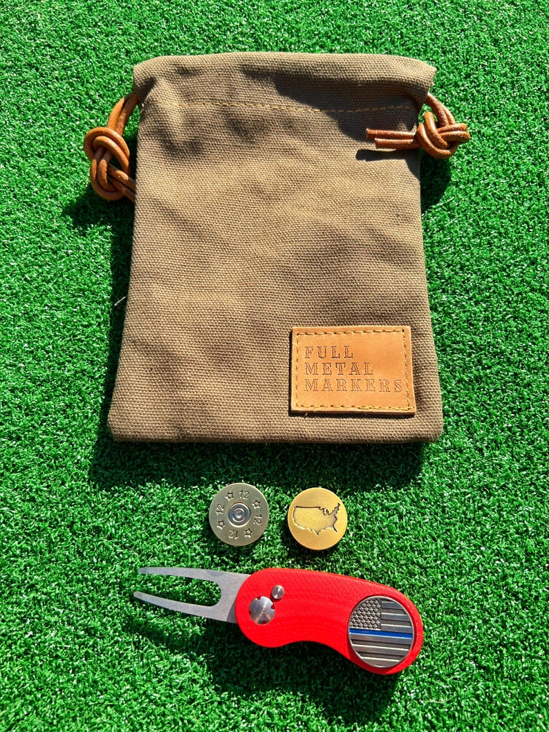 May include: A set of golf accessories including a red golf ball marker with a thin blue line flag design, a gold golf ball marker with a map of the United States, a brass golf ball marker with a bullet design, and a brown canvas bag with a leather patch that reads "Full Metal Markers".