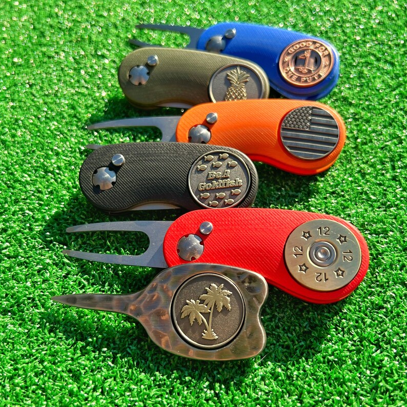 May include: Five different golf divot tools with various designs. The tools are made of metal and have a variety of colors, including black, green, orange, red, and blue. The designs include a pineapple, a fish, a flag, a bullet, and a palm tree.