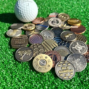 May include: A collection of various golf ball markers, including a golf ball, a pineapple, a palm tree, a bullet, a bear, a fish, and a star. The markers are made of metal and have different designs and colors.