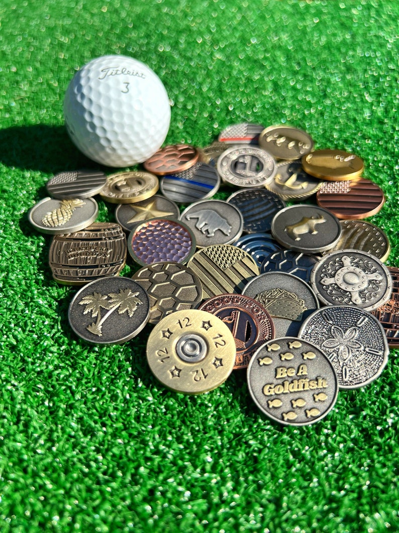 May include: A collection of various golf ball markers, including a golf ball, a pineapple, a palm tree, a bullet, a bear, a fish, and a star. The markers are made of metal and have different designs and colors.