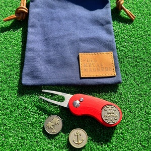 May include: A set of golf accessories including a red golf ball marker with a fish design, a golf ball retriever with a silver fork, and two gold metal golf ball markers with a palm tree and an anchor design. The accessories are on a green artificial grass surface. The golf ball markers are in a blue canvas bag with a brown leather tag that says "Full Metal Markers".