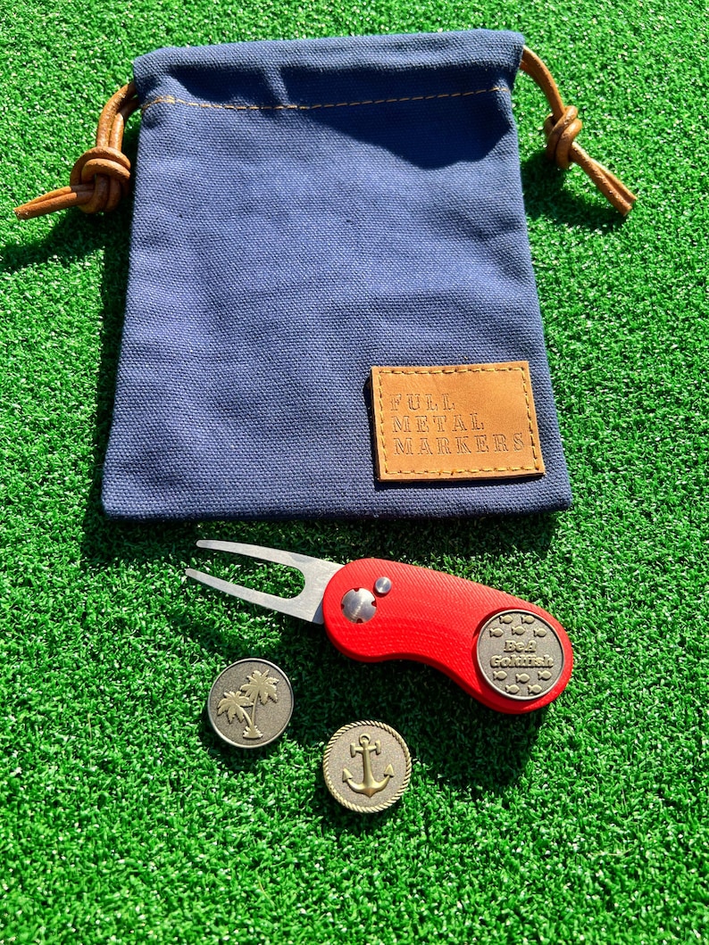 May include: A set of golf accessories including a red golf ball marker with a fish design, a golf ball retriever with a silver fork, and two gold metal golf ball markers with a palm tree and an anchor design. The accessories are on a green artificial grass surface. The golf ball markers are in a blue canvas bag with a brown leather tag that says "Full Metal Markers".