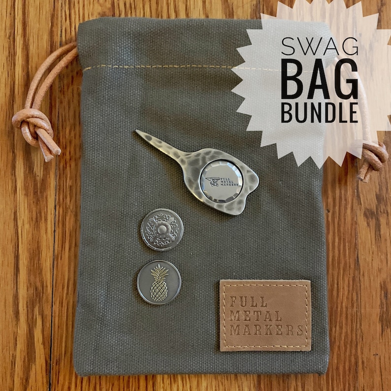 May include: A green canvas bag with a drawstring closure, containing a set of golf accessories. The set includes a divot tool, three metal golf ball markers, and a leather patch that reads "Full Metal Markers".
