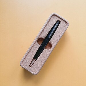 May include: A black pen with a silver tip rests in a beige rectangular case. The pen is centered in the case, which has a textured surface. The background is a solid, light orange color.
