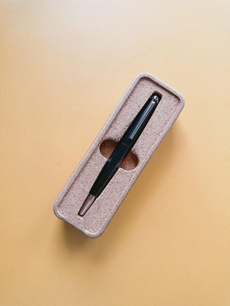 May include: A black pen with a silver tip rests in a beige rectangular case. The pen is centered in the case, which has a textured surface. The background is a solid, light orange color.