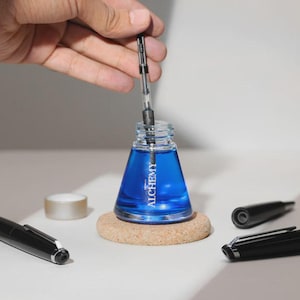 May include: A clear glass ink bottle filled with blue ink, with the word "ALCHEMY" printed on the front. A hand is holding a pen over the bottle. Several black pens and pen parts are on the table, along with a small roll of tape.