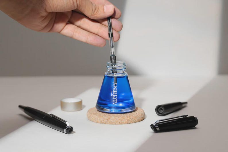 May include: A clear glass ink bottle filled with blue ink, with the word "ALCHEMY" printed on the front. A hand is holding a pen over the bottle. Several black pens and pen parts are on the table, along with a small roll of tape.