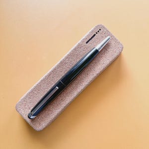 May include: A black fountain pen with a silver tip and clip rests on a rectangular, light brown pen case. The pen case has a textured surface and a small, dark line and dots on the top.