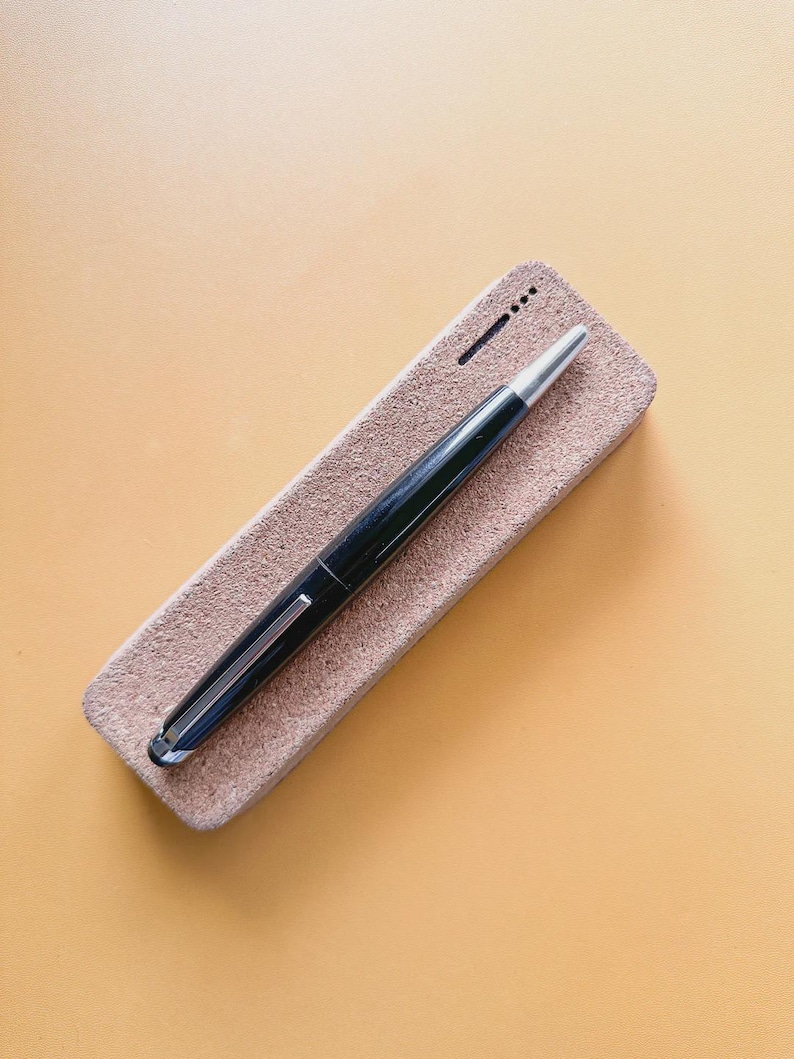 May include: A black fountain pen with a silver tip and clip rests on a rectangular, light brown pen case. The pen case has a textured surface and a small, dark line and dots on the top.