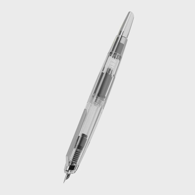 May include: A clear fountain pen with a silver-colored tip and accents. The pen's body is transparent, revealing the internal ink mechanism. The pen is angled diagonally against a plain background.