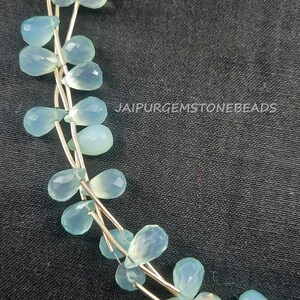 May include: A strand of light blue gemstone beads in a teardrop shape. The beads are strung on a silver wire and are arranged in a loose, flowing pattern. The beads are translucent and have a slight shimmer to them. The image is taken against a dark background.
