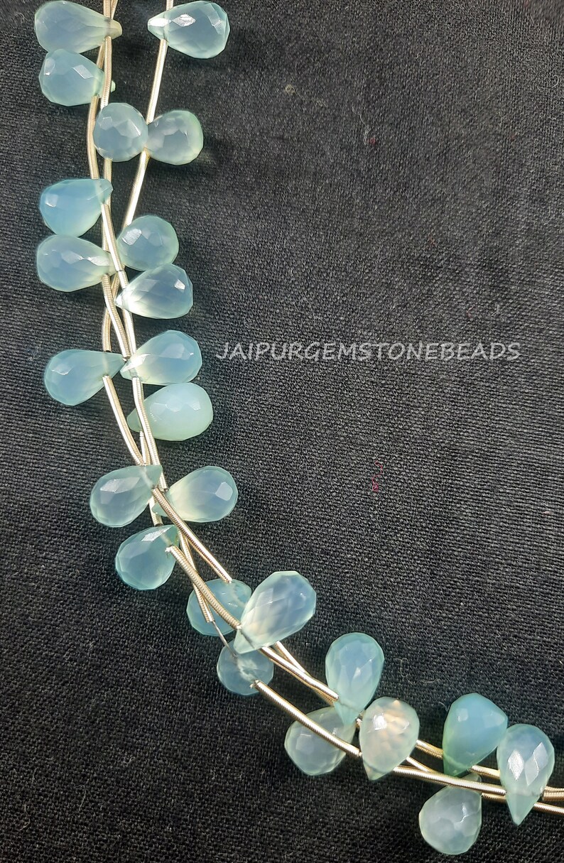 May include: A strand of light blue gemstone beads in a teardrop shape. The beads are strung on a silver wire and are arranged in a loose, flowing pattern. The beads are translucent and have a slight shimmer to them. The image is taken against a dark background.