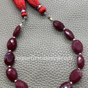 Puede incluir: Un collar de cuentas de piedras preciosas rojo oscuro, ovaladas y facetadas. Las cuentas están ensartadas con espaciadores blancos y tienen borlas rojas en cada extremo. El texto "JaipurGemstoneBeads" es visible.