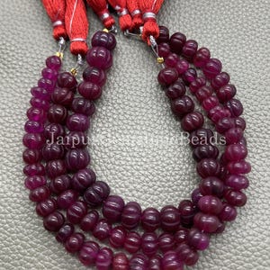 May include: Strands of deep burgundy gemstone beads with a carved pumpkin design. The beads are strung together and attached to red tassels. The text "Jaipur Stone Beads" is visible on the image.
