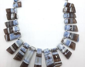 May include: A necklace made of light blue and brown gemstone beads. The beads are faceted and have a unique, striped pattern. The necklace is strung on a silver chain and has a light blue tassel at each end.