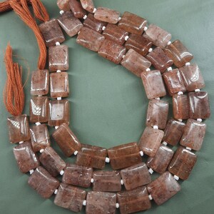 Puede incluir: Una hebra de cuentas de piedras preciosas rectangulares marrones y transparentes con separadores blancos. Las cuentas están enhebradas en un cordón marrón con borlas.