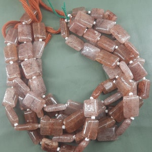 Puede incluir: Una hebra de cuentas de piedra preciosa rectangulares de color marrón y transparente con separadores blancos. Las cuentas están enhebradas en un cordón con borlas marrones.