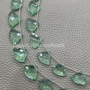 May include: Close-up of a necklace featuring multiple strands of faceted, leaf-shaped, light green gemstones. The gemstones are strung on delicate silver chains. The background is a textured, gray surface. The text "JaipurGemstoneBeads" is visible.