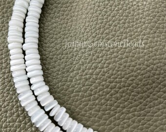 May include: Two strands of white, disc-shaped beads. The beads are strung together, creating a necklace-like appearance. The background is a textured, light brown surface. The beads are uniform in size and shape, reflecting light.