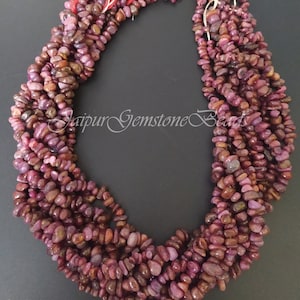 Könnte beinhalten: Eine Strähne von Rubin-Edelsteinen in Chipform. Die Perlen sind tief rotviolett und haben eine natürliche, unregelmäßige Form. Die Perlen sind auf einem dünnen Faden aufgefädelt und bereit für die Schmuckherstellung.