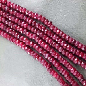 Puede incluir: Una hebra de cuentas de rub&iacute; rojo intenso, facetadas y pulidas, dispuestas en filas sobre un fondo gris. Las cuentas son uniformes en tama&ntilde;o y forma, creando un patr&oacute;n visualmente atractivo.