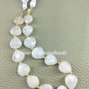 May include: A strand of heart-shaped, iridescent moonstone beads with gold-colored spacers. The beads are strung together with white tassels at the top. The text "JaipurGemstoneBeads" is visible on the image.
