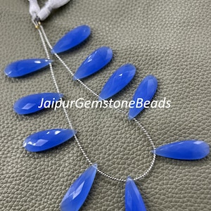 Könnte beinhalten: Eine Kette aus facettierten, tropfenförmigen blauen Edelsteinperlen. Die Perlen sind auf einer silbernen Kette aufgereiht und werden vor einem strukturierten, graugrünen Hintergrund präsentiert. Der Text "JaipurGemstoneBeads" ist sichtbar.