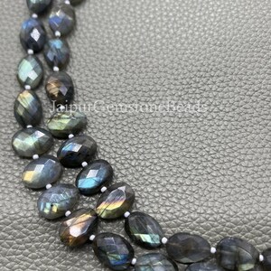 Puede incluir: Primer plano de un collar hecho de cuentas facetadas de piedra labradorita. Las cuentas ovaladas muestran colores iridiscentes de azul, verde y dorado. Las cuentas están ensartadas en un hilo blanco.