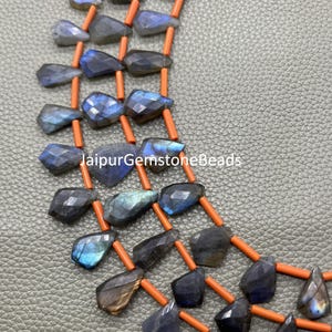 May include: Close-up of a necklace featuring faceted labradorite gemstones in shades of blue, gray, and brown. The gemstones are strung on orange-colored strands. The text "jaipurgemstonebeads" is visible.