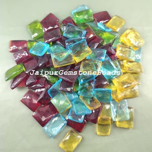 Puede incluir: Surtido de gemas de vidrio rectangulares facetadas y de colores. Las gemas son en tonos de rojo, verde, azul y amarillo. JaipurGemstoneBeads está impreso en la imagen.