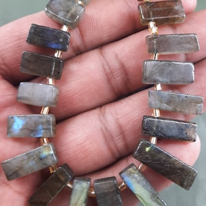 Puede incluir: Una hebra de cuentas de piedra preciosa de labradorita gris oscuro con destellos iridiscentes azules y verdes. Las cuentas son rectangulares y tienen un acabado liso y pulido.