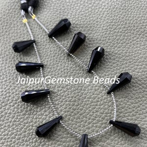 May include: A strand of black gemstone beads, teardrop-shaped and strung on a silver chain. The beads are faceted and glossy. The image includes a black tassel and the text "JaipurGemstone Beads".