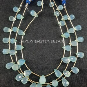 May include: A strand of light blue gemstone beads in a teardrop shape. The beads are strung on a silver wire and are approximately 10mm in size. The beads are arranged in a repeating pattern of three beads per row. The image includes the text "JAIPURGEMSTONEBEADS".