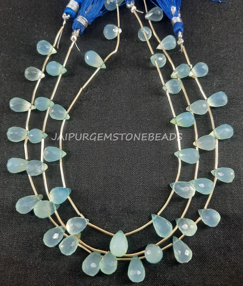 May include: A strand of light blue gemstone beads in a teardrop shape. The beads are strung on a silver wire and are approximately 10mm in size. The beads are arranged in a repeating pattern of three beads per row. The image includes the text "JAIPURGEMSTONEBEADS".