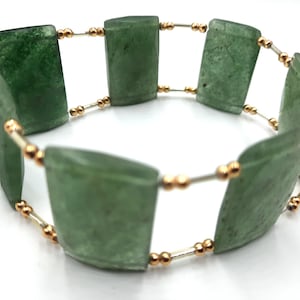 Puede incluir: Un brazalete de piedras preciosas verdes con cuentas doradas y alambre plateado. El brazalete está hecho de piedras de forma rectangular.