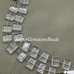 May include: Clear, rectangular gemstone beads strung on a silver wire, with white tassels at the ends. The beads are arranged in strands against a textured, olive-green surface. The text "Jaipur Gemstone Beads" is visible.