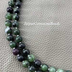 May include: Close-up of two strands of gemstone beads. The beads are a mix of green, black, and burgundy colors. The text "JaipurGemstoneBeads" is visible in the background. The beads are strung on a white string.