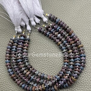 Puede incluir: Cuatro hilos de cuentas de piedras preciosas en forma de disco en tonos azul, morado y marrón, separadas por pequeños espaciadores blancos. Cada hilo termina con una borla de hilo blanco. El texto JaipurGemstoneBeads es visible.