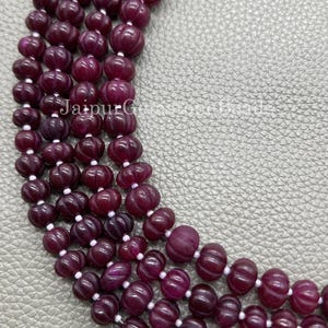 May include: Close-up of several strands of deep burgundy gemstone beads. The beads have a carved, textured design and are strung on white string. The text "Jaipur Gemstone Beads" is visible in the image.