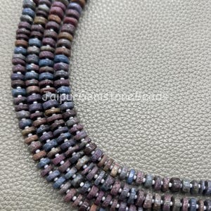 May include: Four strands of small, disc-shaped beads in muted shades of blue, purple, and brown. The beads are strung on a thin white cord, creating a textured, multi-strand arrangement. The text JaipurGemstoneBeads is visible in the centre.