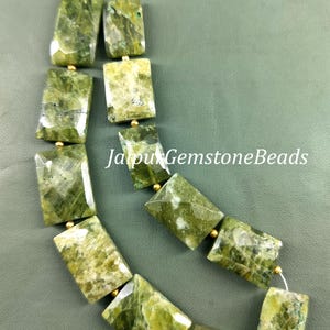 Puede incluir: Una hebra de cuentas de piedras preciosas verdes con detalles dorados. Las cuentas son rectangulares y tienen un aspecto moteado y nublado. La hebra se muestra sobre una superficie verde. "Jaipur Gemstone Beads" está escrito en blanco sobre la superficie.