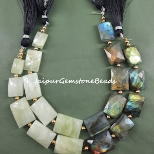 Puede incluir: Una hebra de cuentas de piedras preciosas con una mezcla de piedras verdes y azul verdoso iridiscente. Las cuentas son facetadas y tienen forma rectangular. La hebra está acabada con borlas negras.