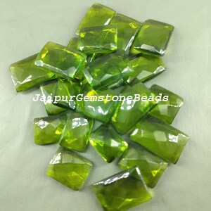 Puede incluir: Un montón de 18 cuentas de piedras preciosas verdes, cada una con una forma rectangular facetada. Las cuentas son translúcidas y tienen un ligero tono amarillo. Las cuentas están dispuestas en un montón aleatorio, con algunas superpuestas. El texto "JaipurGemstoneBeads" es visible en la imagen.